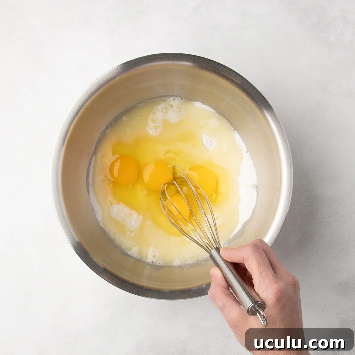 Hand holding a metal whisk about to whisk together a mixture of white dry ingredients with eggs and lemon juice liquid mixture.