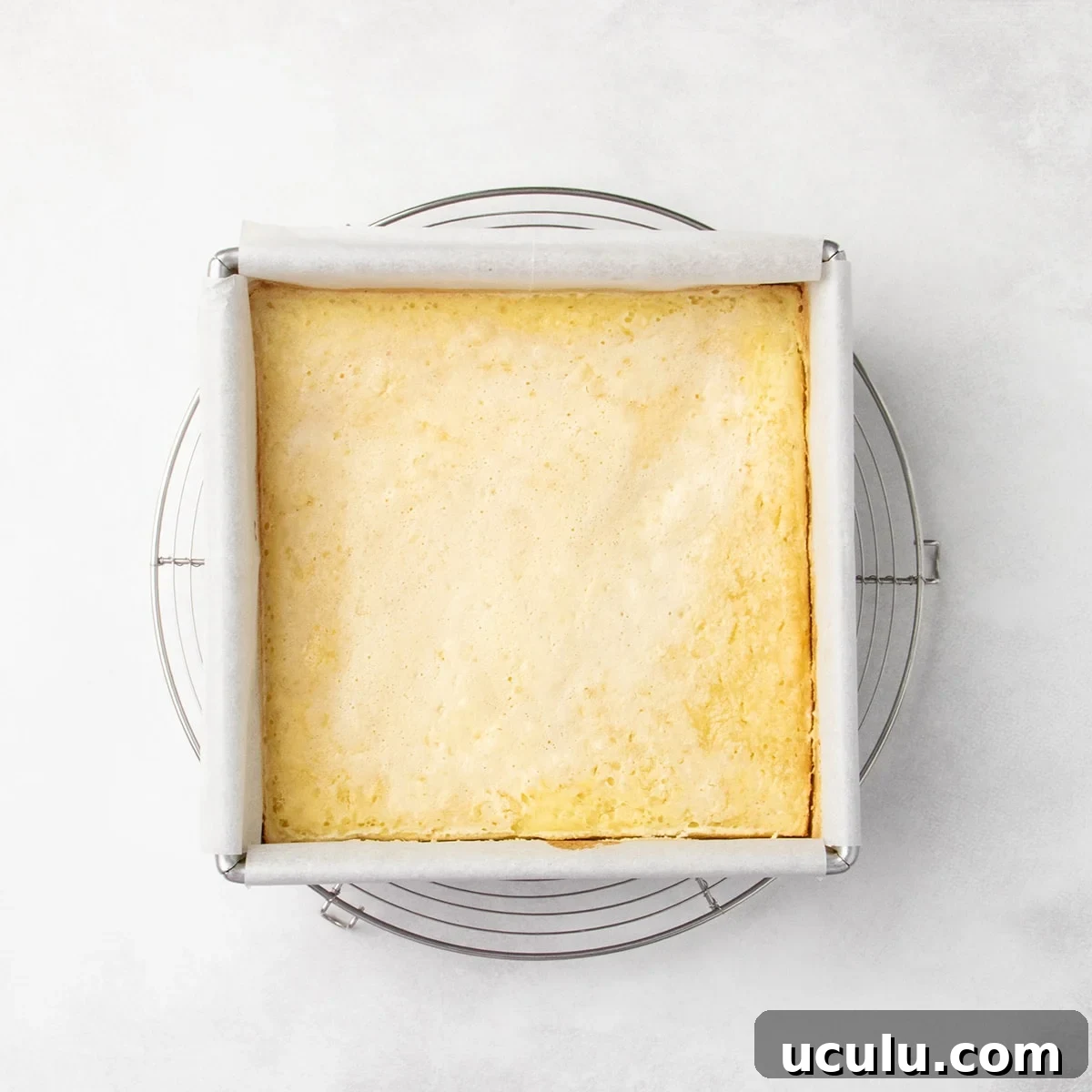 Uncut, baked lemon bars in a parchment-lined square pan resting on a round wire rack, lightly golden around the edges.