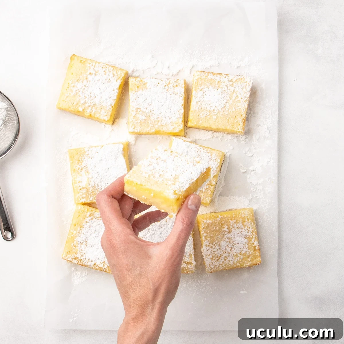 Hand holding a square lemon bar dusted with powdered sugar over several cut bars on parchment paper, with a sieve partially visible to the side.