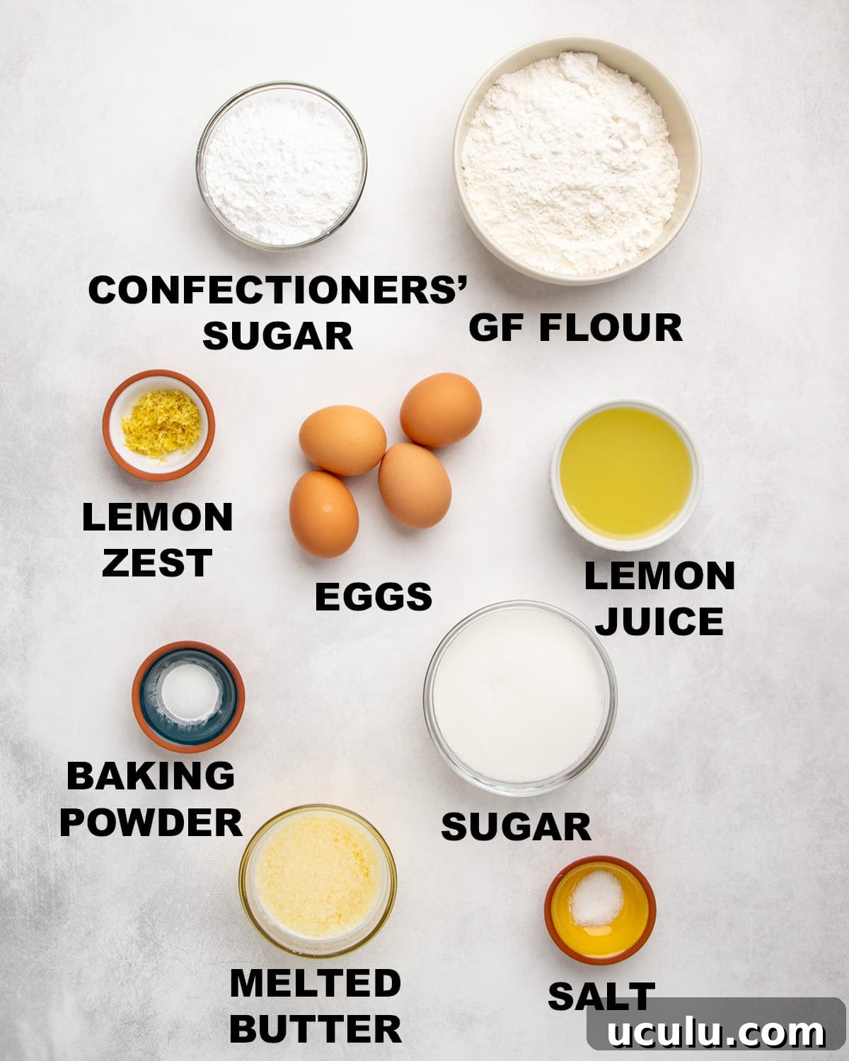 Overhead view of measured ingredients in bowls with labels including confectioners’ sugar, gluten free flour, lemon zest, eggs, lemon juice, baking powder, sugar, melted butter, and salt.