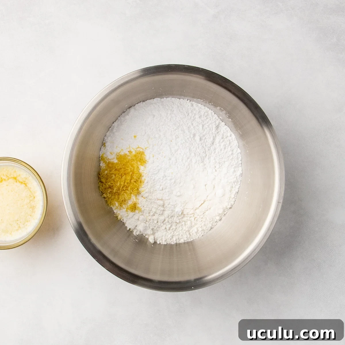 Metal mixing bowl filled with white dry ingredients and a small pile of yellow lemon zest, with a glass of melted butter mixture partially visible to the side.
