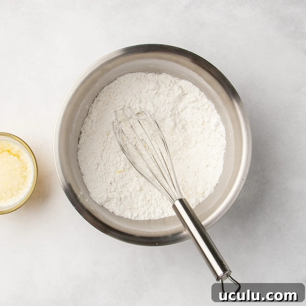 Metal mixing bowl with white dry ingredients being whisked with a metal whisk, with some visible yellow flecks of lemon zest