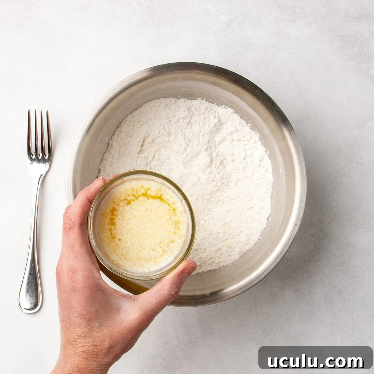 Hand holding a small glass of melted butter mixture about to pour it into a bowl of white dry ingredients, with a fork resting nearby.
