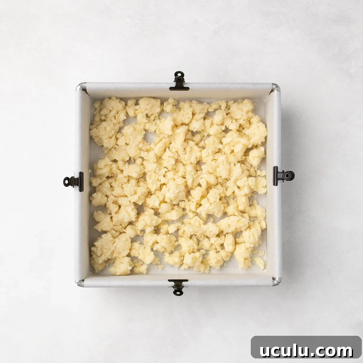 Pale dough clumps scattered in an even layer in a square baking pan lined with parchment paper, clipped to each of the four sides with a small black binder clip.