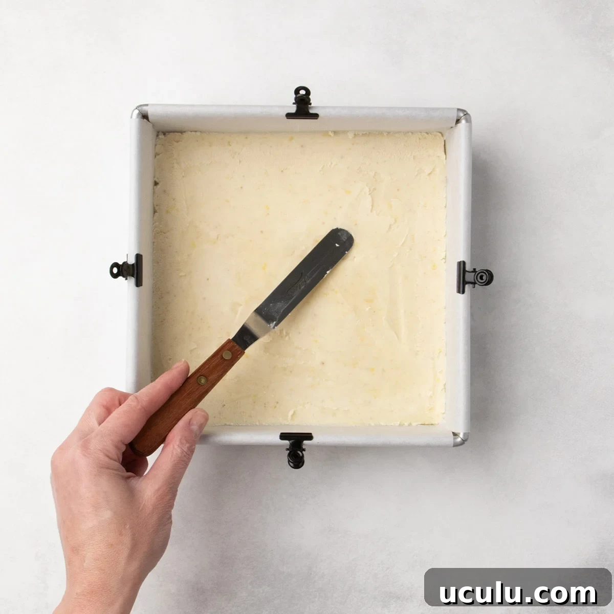 Hand holding a small offset spatula to press pale dough into a smooth, even layer in a parchment-lined square pan.