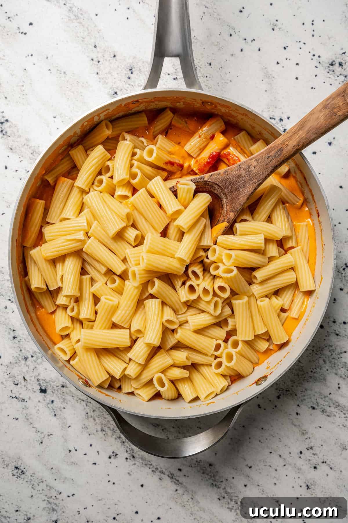 Rigatoni pasta added to vodka sauce in a large skillet, with a wooden spoon.