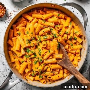 Overhead view of spicy rigatoni alla vodka garnished with chopped parsley in a large skillet, with a wooden spoon and small bowls of garnishes on the side.