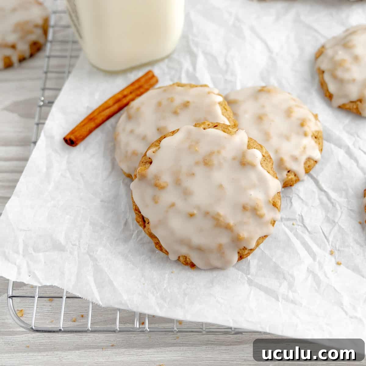 Three oatmeal cookies on parchment paper. Milk, cookies and a cinnamon stick in the background.