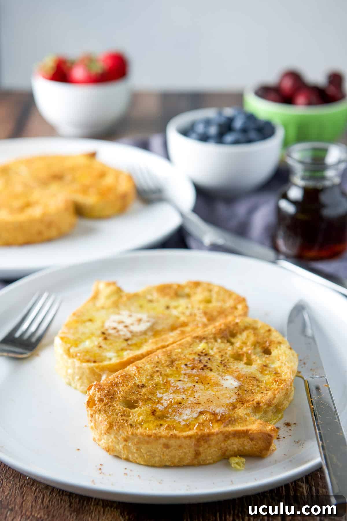 Plates with the French toast with melted butter and cinnamon sprinkled; fruit and maple syrup in back