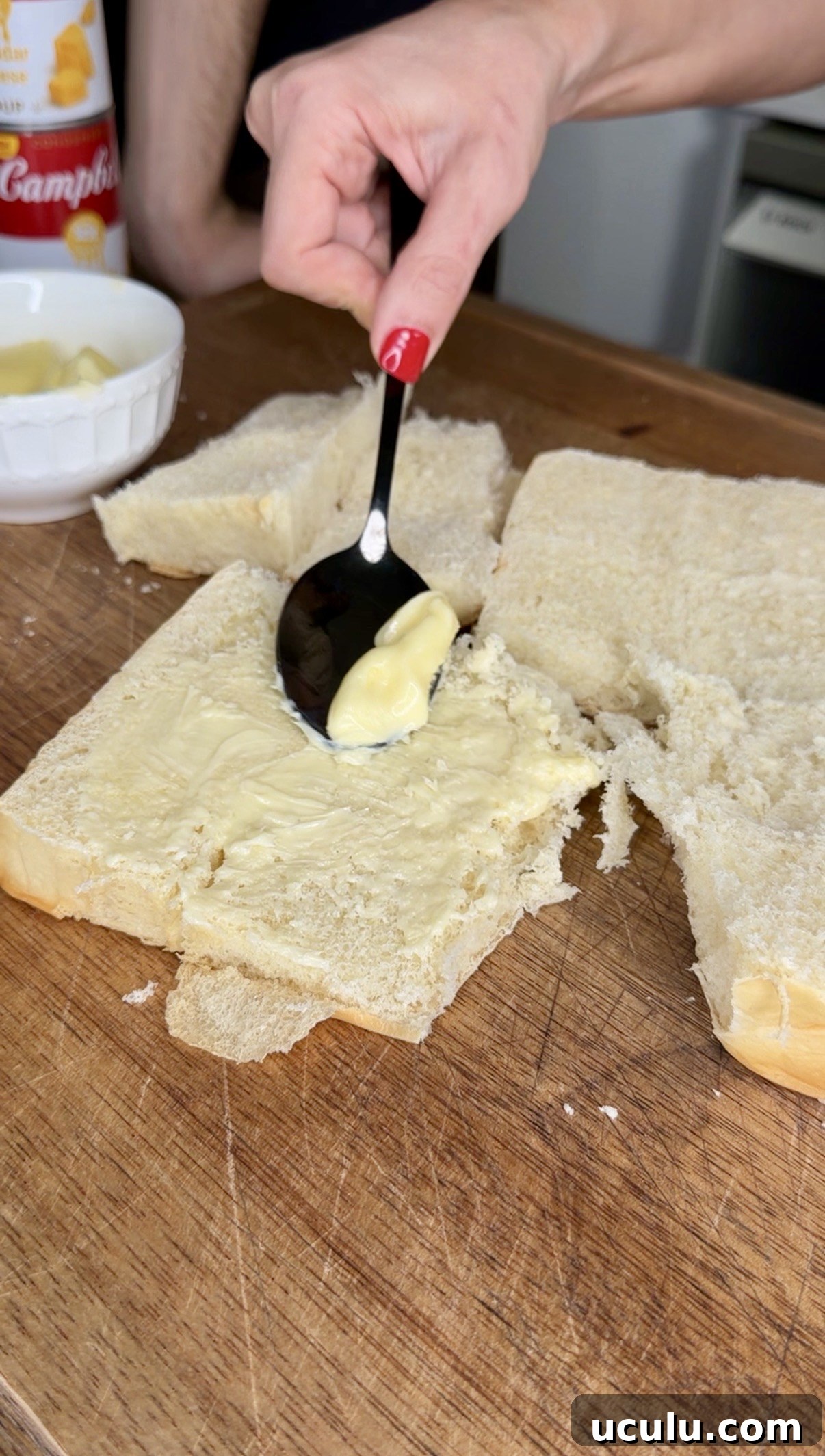 A hand with red nail polish spreads butter onto a slice of white bread for a Cheddar Make-Ahead Breakfast casserole, using a spoon on a wooden cutting board, with extra bread slices and a bowl of butter nearby.