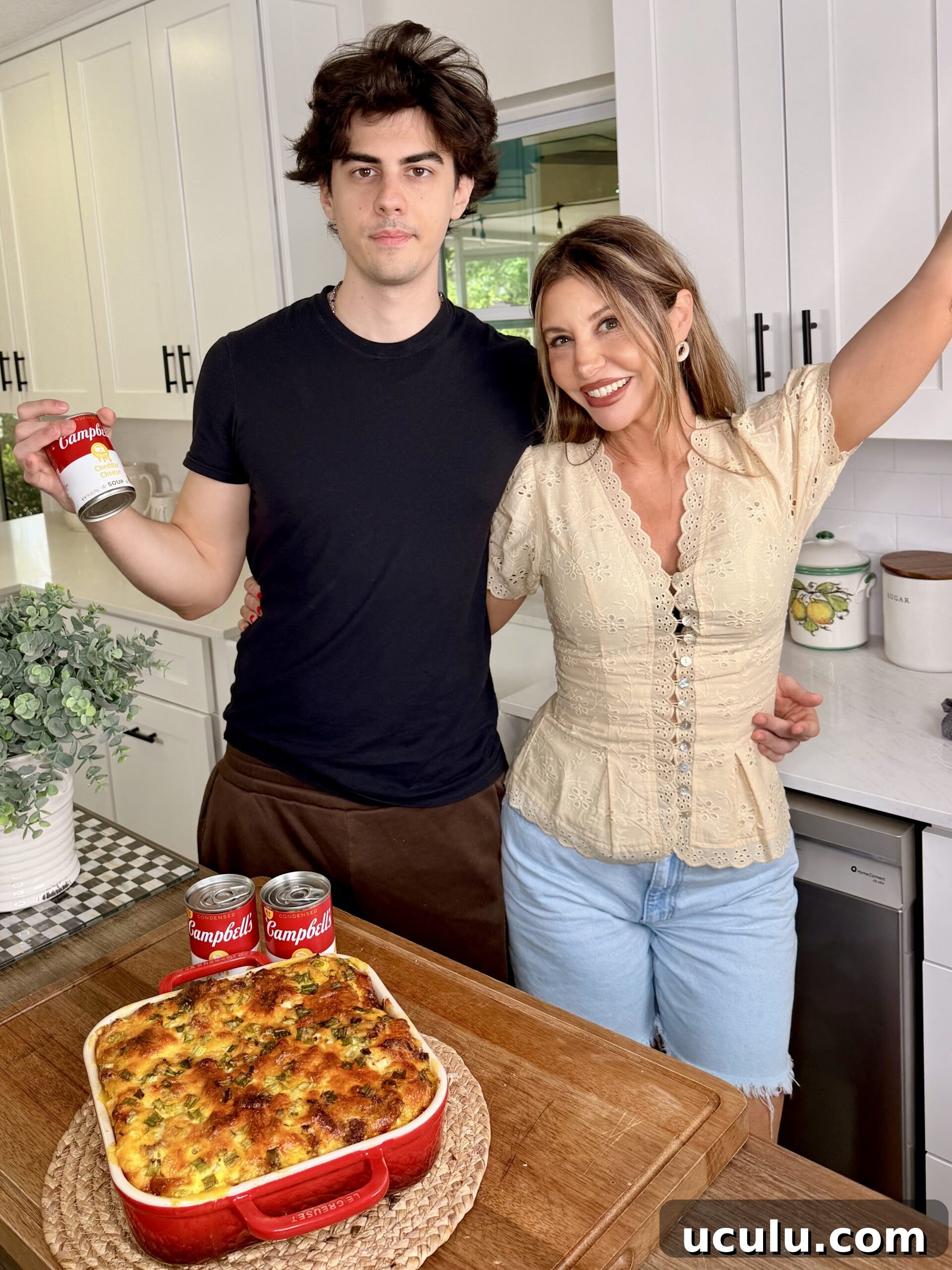 A young man and a woman stand in a kitchen, smiling beside a Cheddar Make-Ahead Breakfast casserole in a red dish. The man holds a can of Campbell’s soup, with two more cans placed on the counter in front of the casserole.