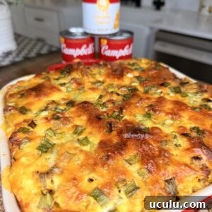 A Cheddar Make-Ahead Breakfast casserole topped with melted cheese and green onions sits in a red dish. Behind it are three cans of Campbell’s soup, one labeled “Cheddar Cheese,” set in a bright kitchen.