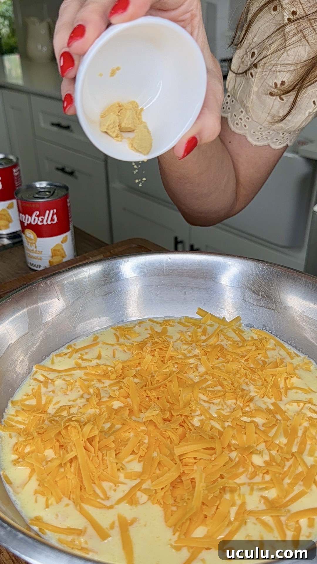A person sprinkles a powdered ingredient from a small white bowl into a pan of eggs and shredded cheddar cheese, preparing a Cheddar Make-Ahead Breakfast casserole, with Campbell’s soup cans visible in the background on the kitchen counter.