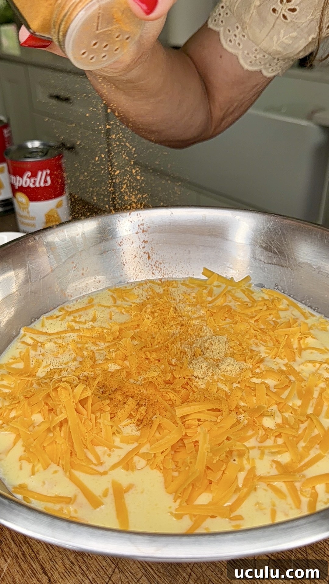 A woman's hand sprinkles seasoning into a large metal bowl filled with shredded cheddar cheese and creamy liquid—the perfect start for a Cheddar Make-Ahead Breakfast casserole. In the background, a can of Campbell's soup sits on the kitchen counter.