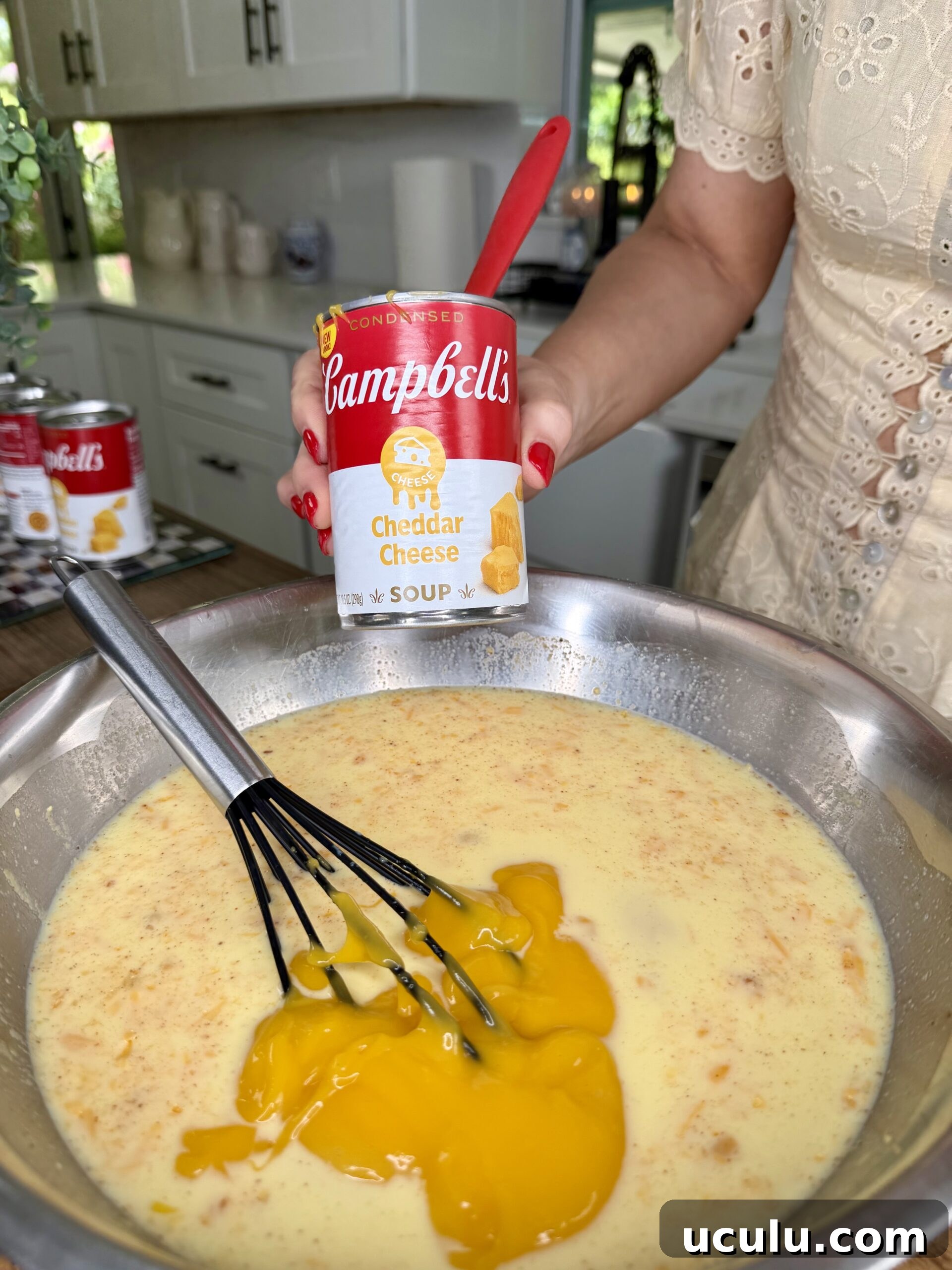 A person holds a can of Campbell’s condensed cheddar cheese soup above a metal bowl filled with a yellow mixture being whisked, preparing ingredients for a Cheddar Make-Ahead Breakfast casserole in a bright kitchen setting.