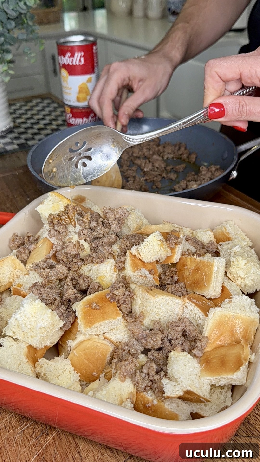 A person prepares a Cheddar Make-Ahead Breakfast casserole by spooning cooked ground beef over cubed bread in a baking dish, with a can of Campbell's soup and a skillet of beef in the background. Another hand steadies the pan.
