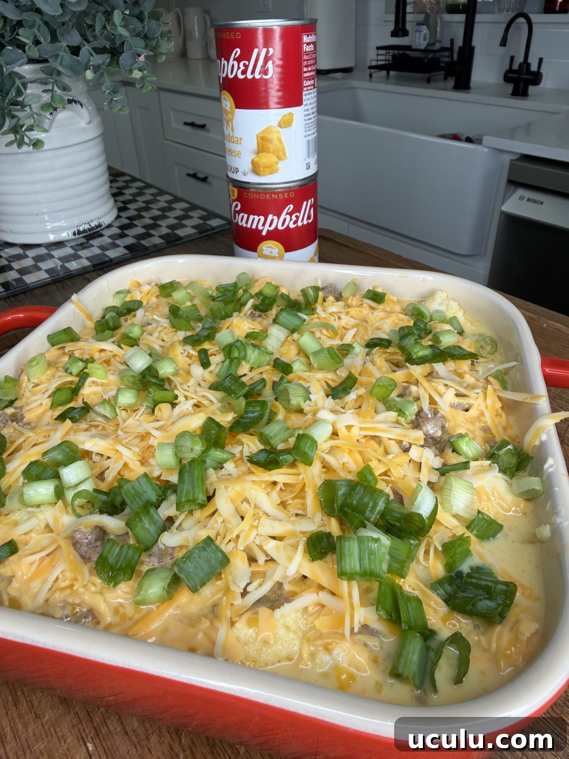 A red baking dish filled with a Cheddar Make-Ahead Breakfast casserole topped with chopped green onions sits on a kitchen counter. Behind it are two stacked cans of Campbell’s soup and part of a modern kitchen is visible in the background.