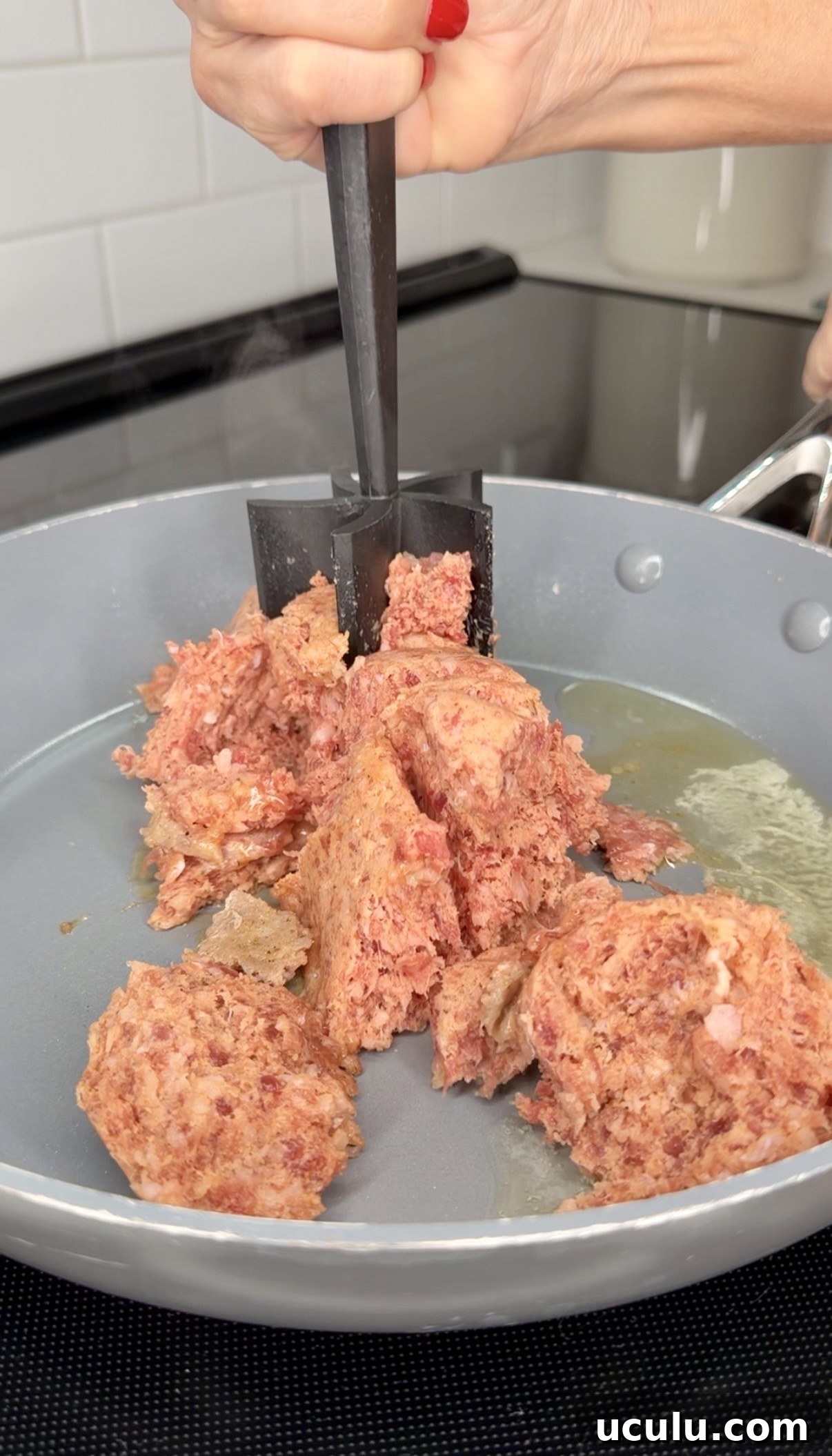 A person uses a black spatula to break up raw ground meat in a gray frying pan on a stovetop, likely preparing ingredients for a Cheddar Make-Ahead Breakfast casserole. Oil is visible in the pan.