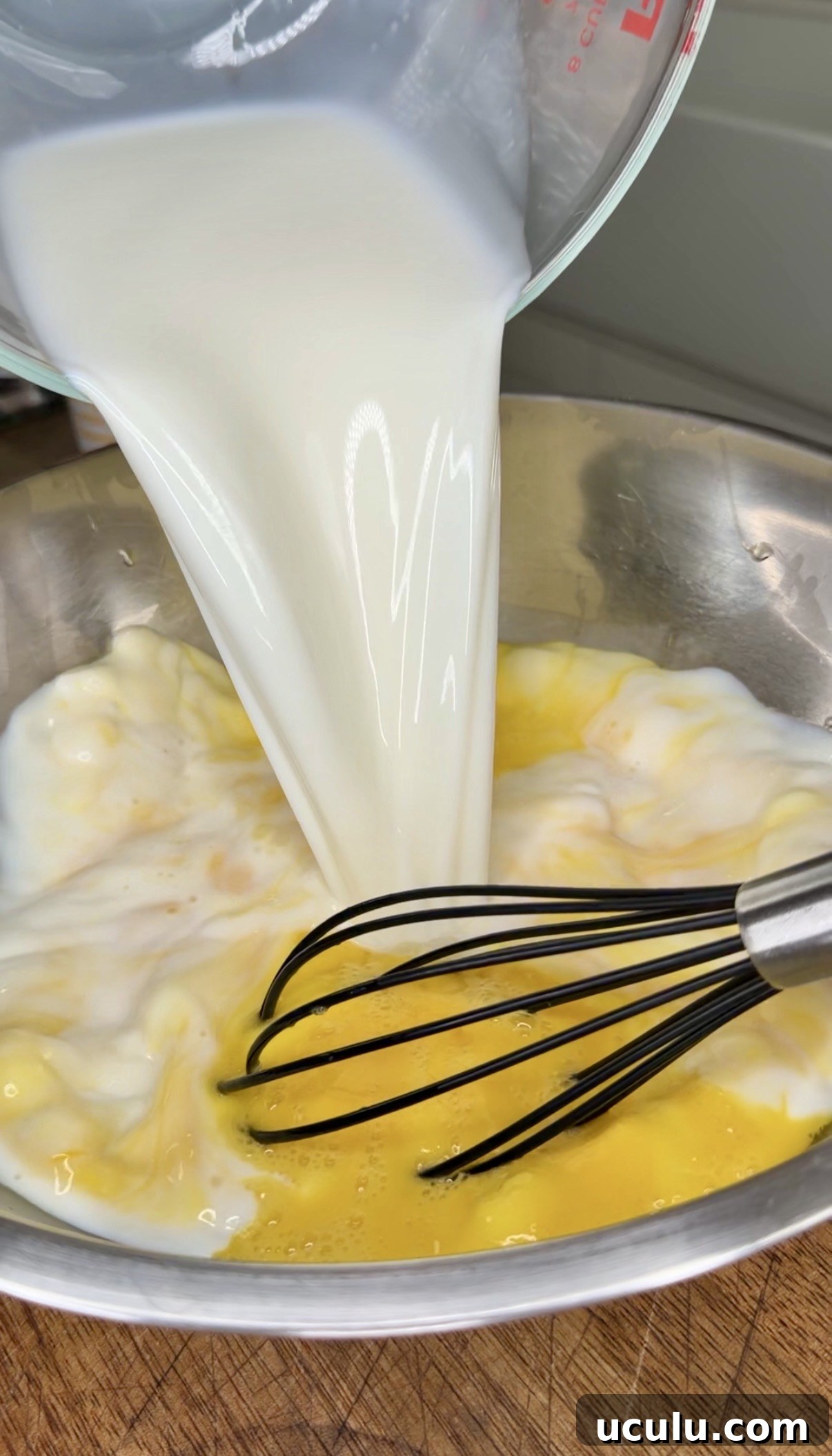 Milk is being poured from a measuring cup into a metal bowl containing beaten eggs for a Cheddar Make-Ahead Breakfast casserole, with a whisk resting inside the bowl.
