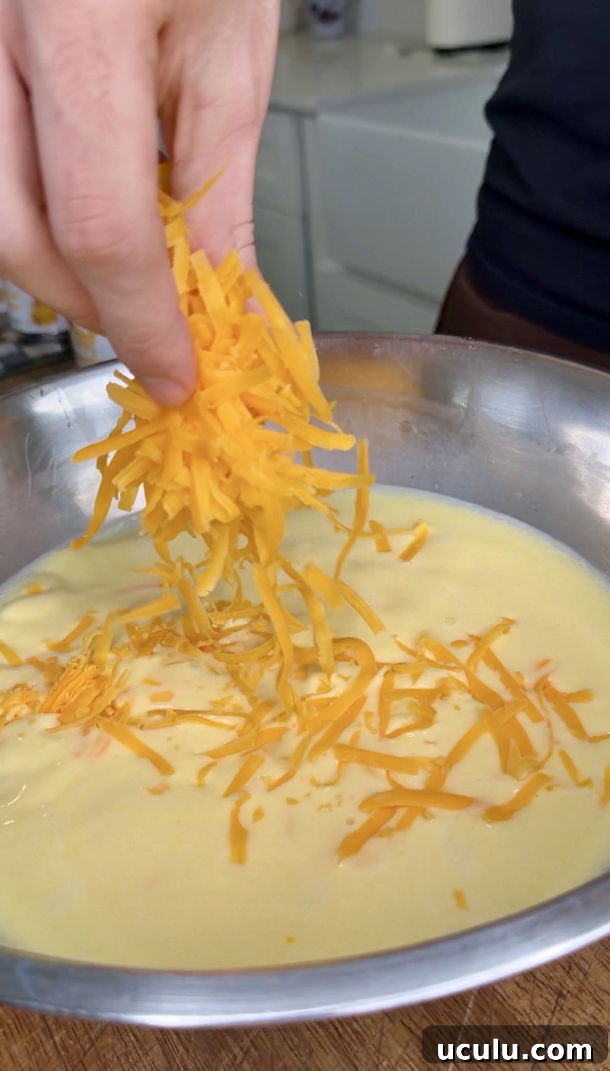A hand sprinkles shredded cheddar into a bowl of creamy yellow mixture, perfect for preparing a Cheddar Make-Ahead Breakfast casserole.