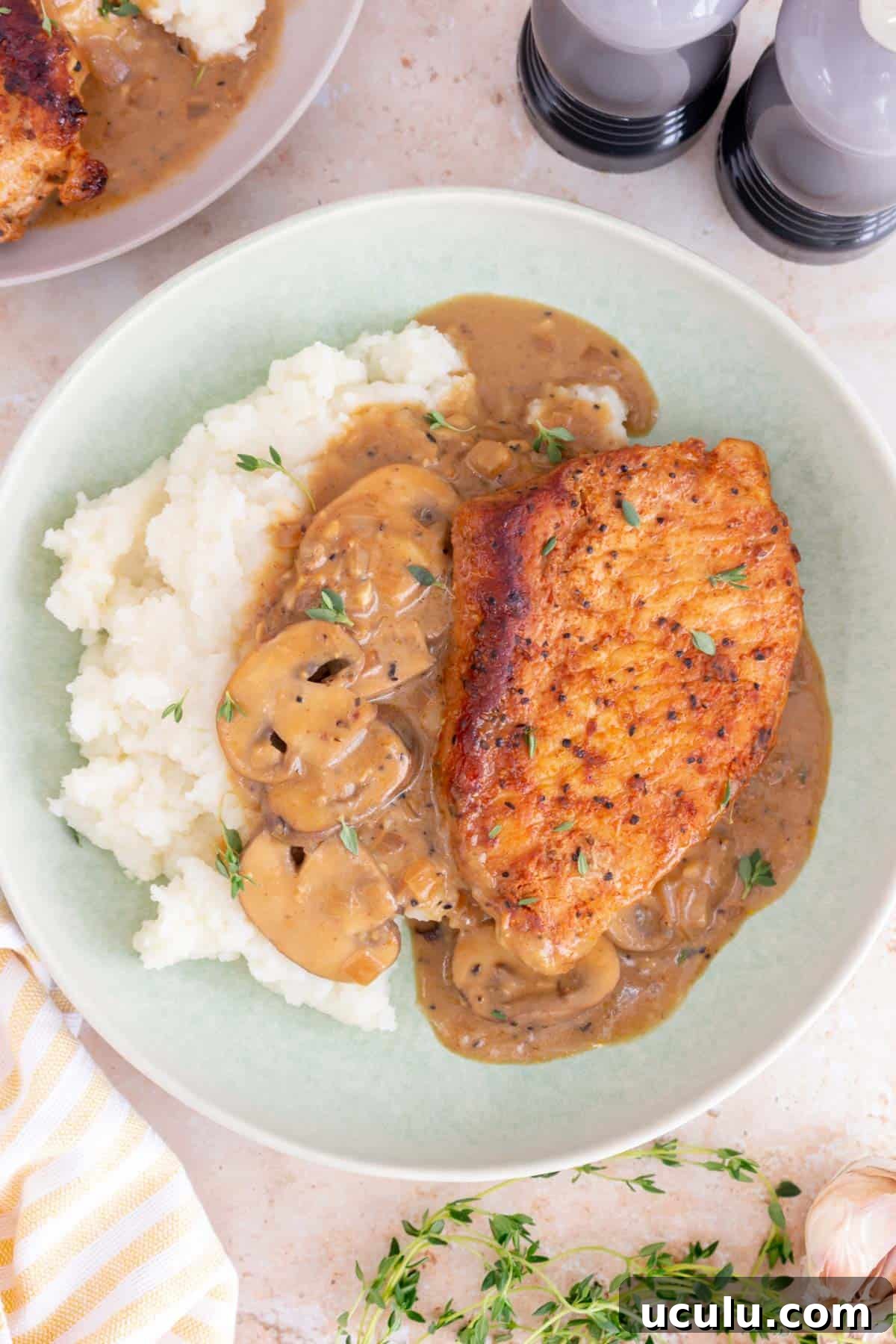 A plate of mashed potatoes with a dutch oven pork chop, sauce, mushrooms, and thyme.