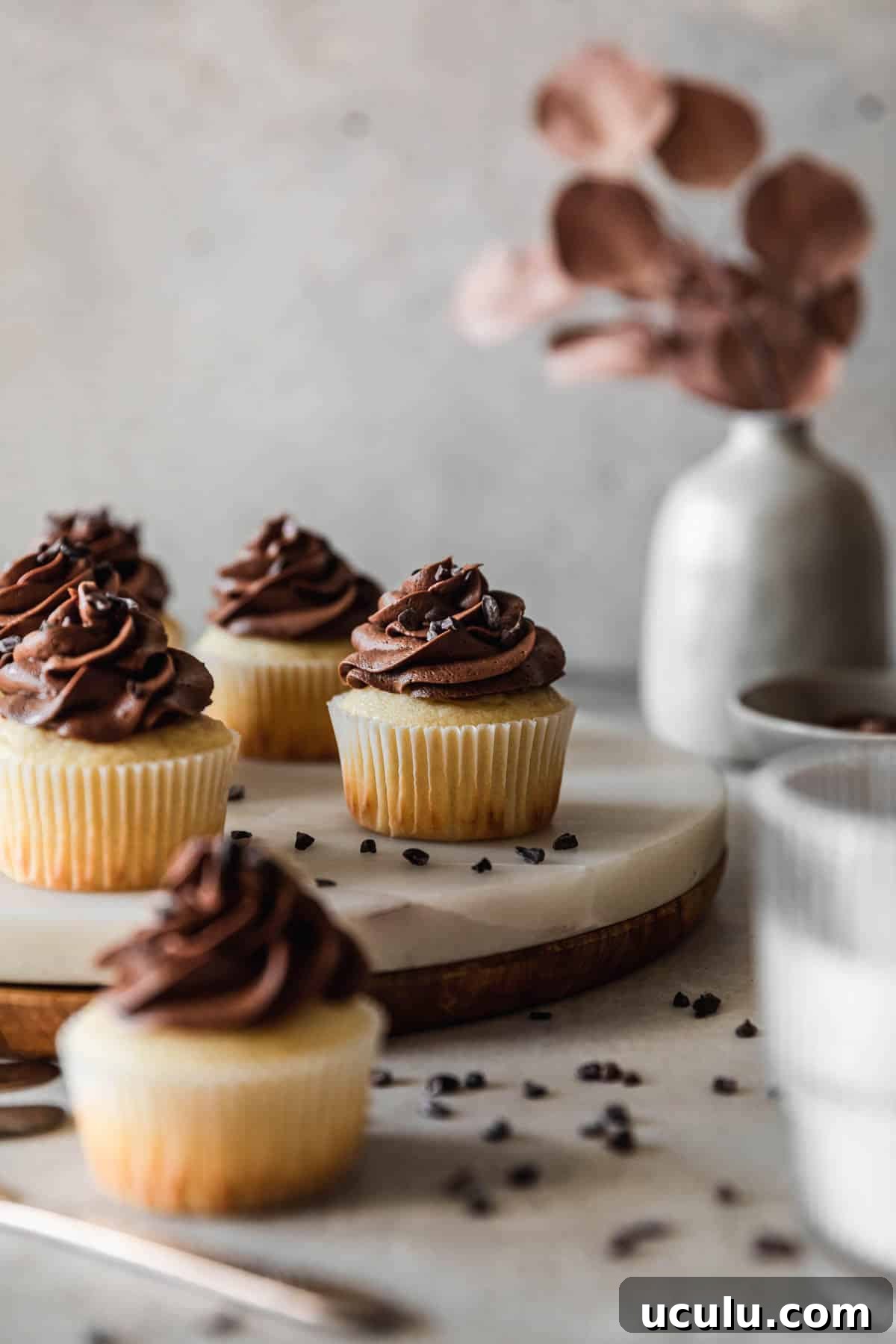 A closeup side image of a marble and wood board topped with high altitude yellow cupcakes with chocolate buttercream on a grey table next to a glass of milk, grey bowl of frosting, and grey vase with dried eucalyptus leaves.