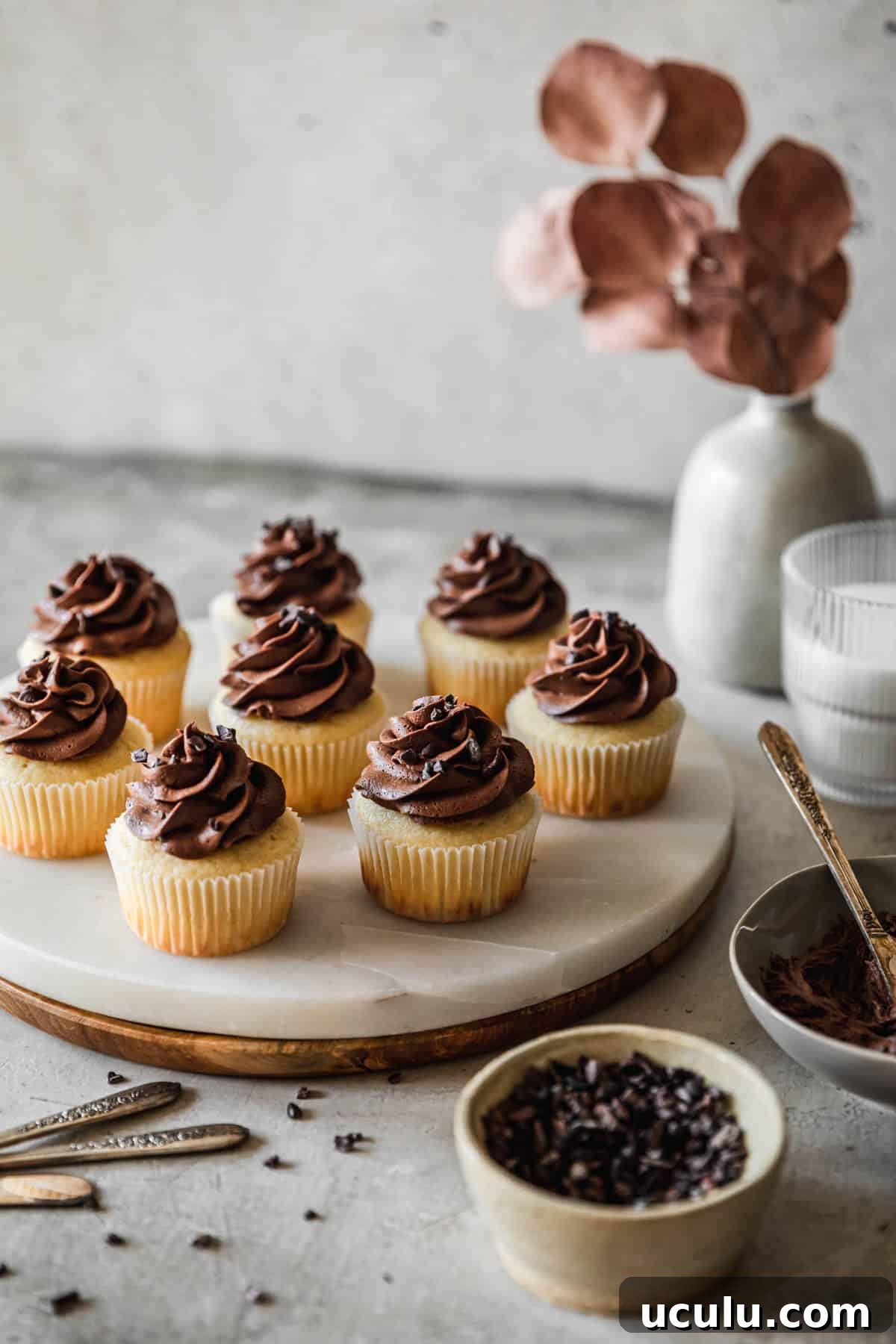 A side image of a marble and wood tray of high altitude yellow cupcakes with chocolate buttercream on a grey table next to a grey bowl of frosting, white bowl of cacao nibs, white vase with brown blossoms, and cup of milk.