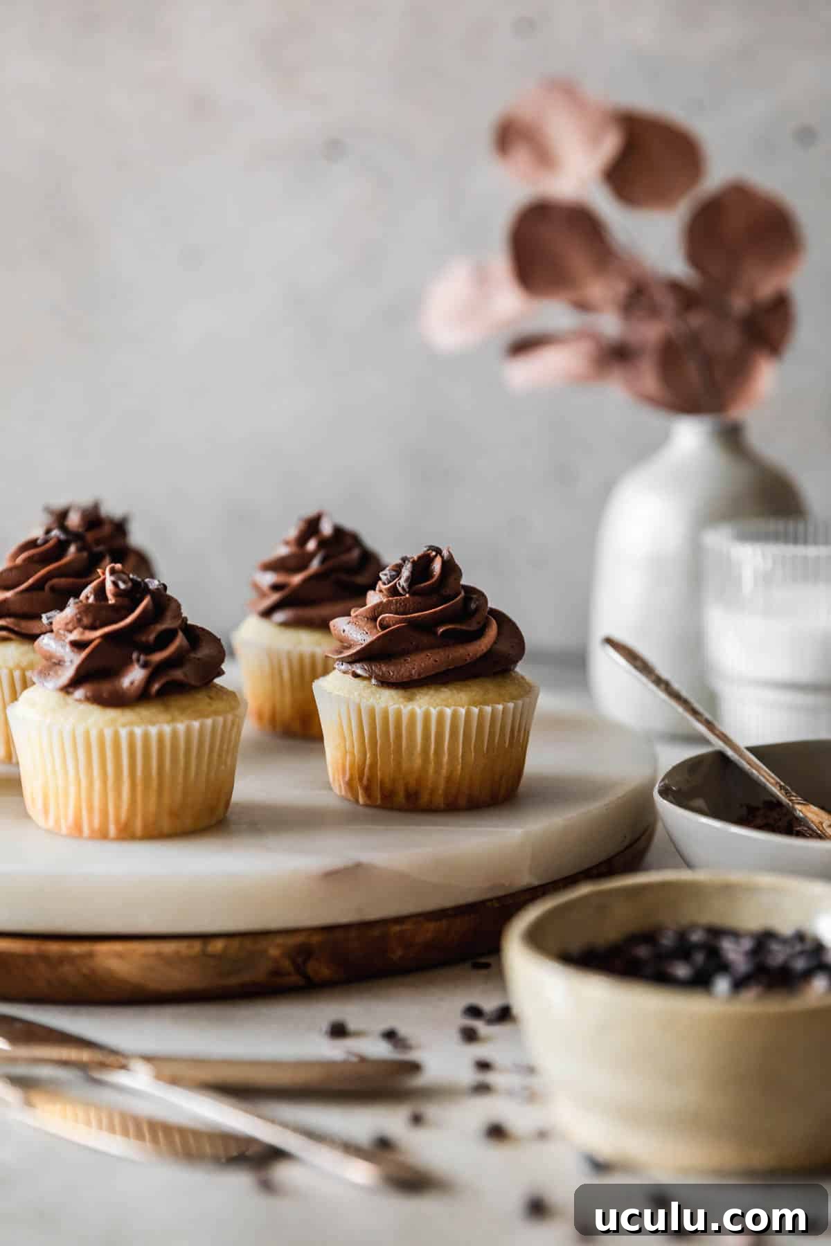 A closeup side image of high altitude yellow cupcakes with chocolate buttercream on a marble and wood cake stand next to a grey vase with blossoms, a glass of milk, a grey bowl of frosting, and a white bowl of cacao nibs with a grey background.