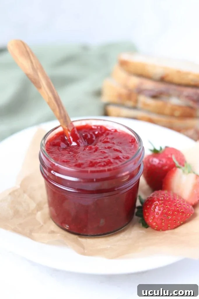 A small jar of strawberry jam sitting on a white plate alongside some fresh strawberries. There is a wooden spoon in the jar and some sourdough in the background.