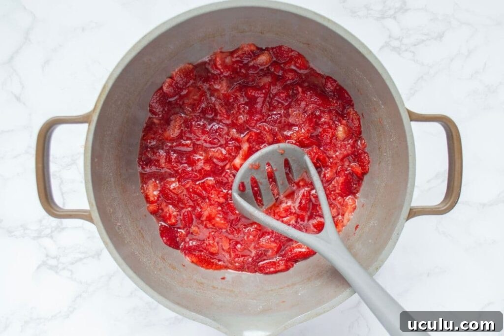 A large handled pot containing mashed red strawberries. There is a potato masher in the pot.