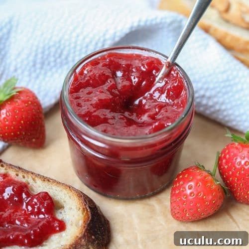 A small jar of strawberry jam with a spoon in it. There are fresh strawberries scattered around it.