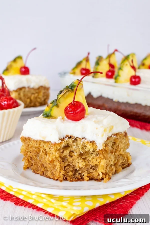 A white plate with a piece of pineapple sunshine cake topped with pineapple cream cheese frosting, a cherry, and pineapple slice on it