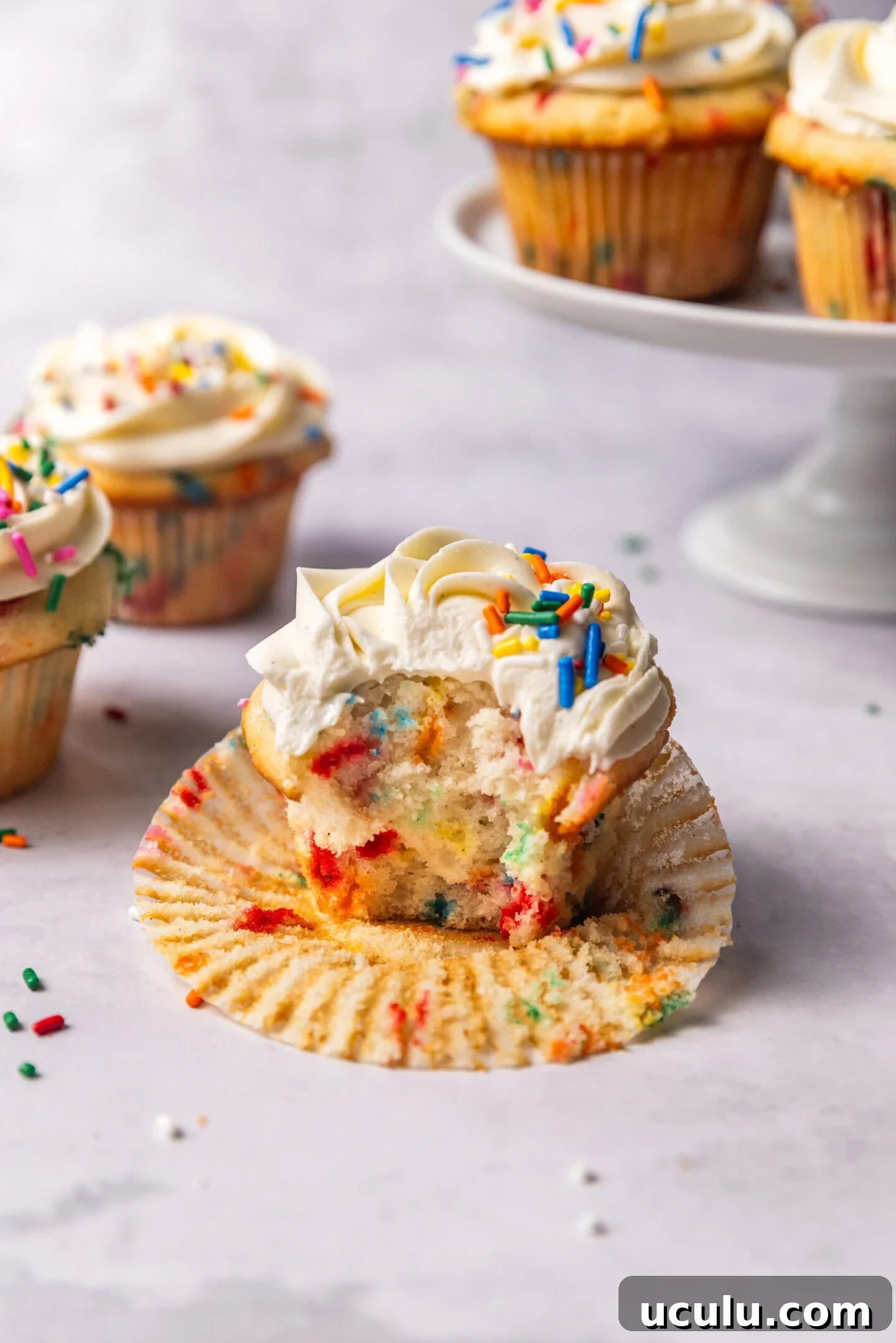 A bite taken out of a cupcake showing the moist, soft and fluffy interior with pops of color from the rainbow sprinkles. Other funfetti cupcakes are sitting in the background on top of a cake stand.