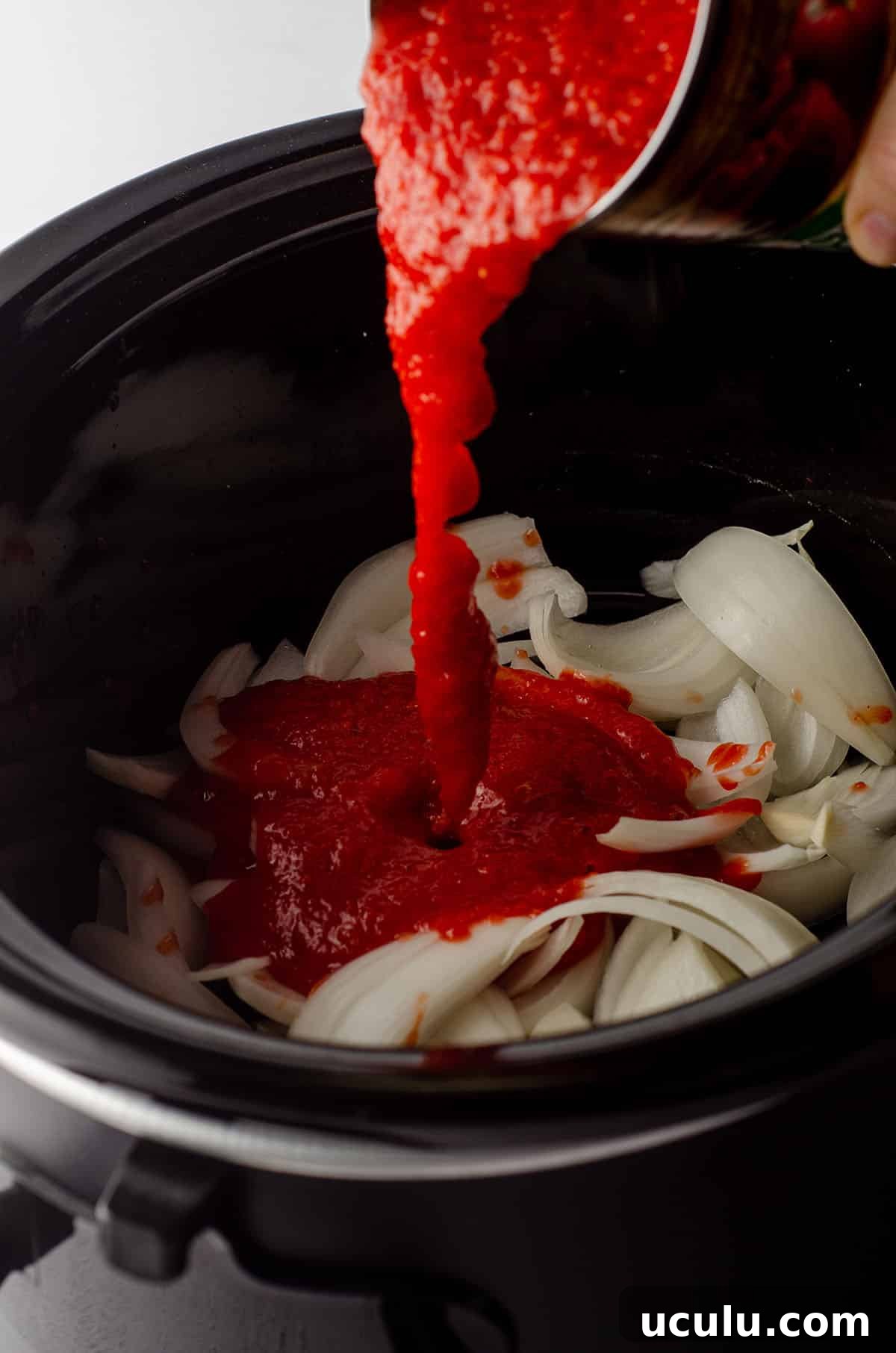 slow cooker filled with sliced onions and a can of crushed tomatoes being poured on top