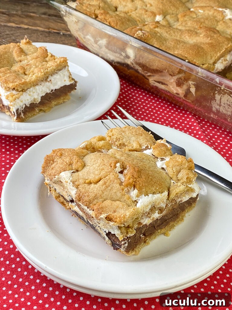 Pieces of smore cake on white plates with layers of graham cracker crust, marshmallows and chocolate bars resting on a crust of graham cracker crumbs on white plates with a fork in the background on a red napkin with small white polka dots