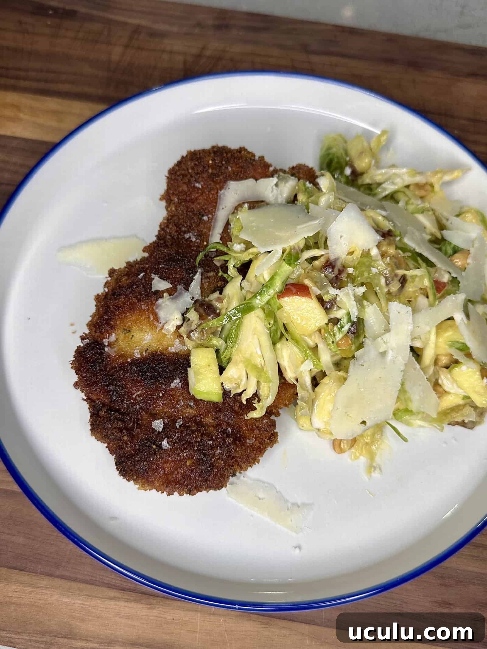 A white plate with crispy breaded cutlets and a mixed salad topped with shaved cheese, set on a wooden surface.