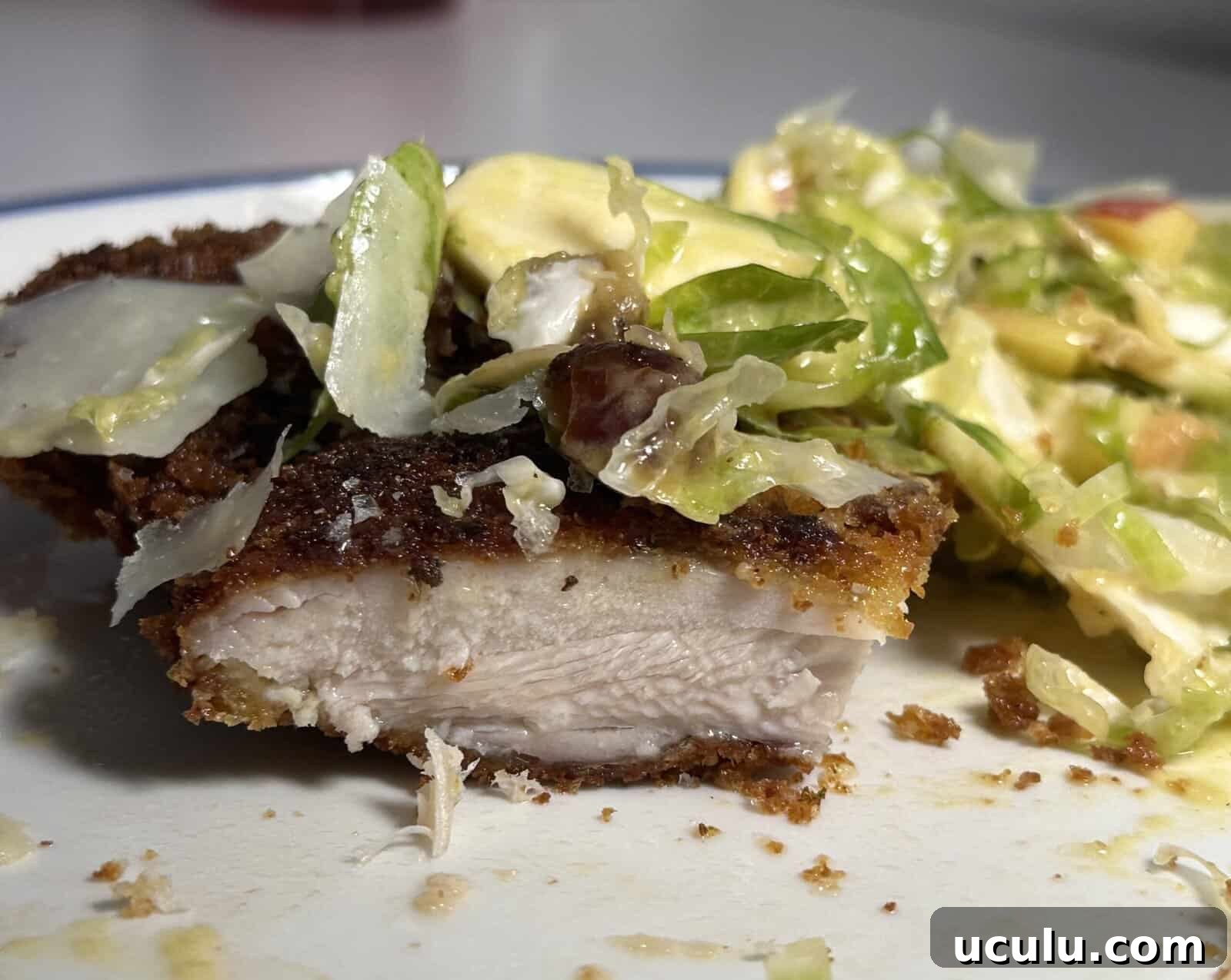 Close-up of a plate with a piece of breaded, cooked chicken cut in half, showing the white interior, next to a creamy salad with lettuce, avocado, and bacon.