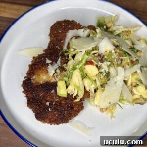 A white plate with crispy breaded cutlets and a mixed salad topped with shaved cheese, set on a wooden surface.