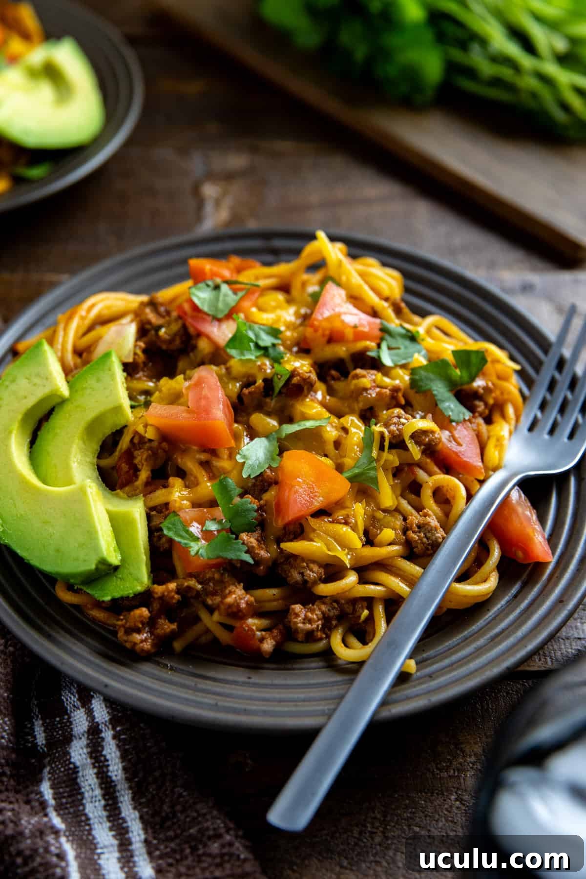 Mexican spaghetti topped with chopped tomatoes and fresh cilantro on brown plate with fork on the side.  Avocado slices on the side.