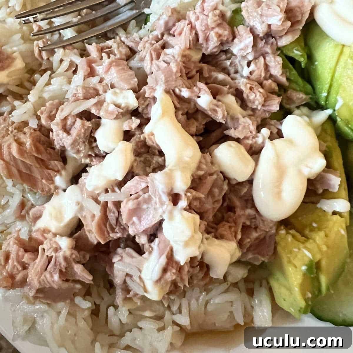 A colorful tuna rice bowl topped with sliced avocado and fresh cucumber on a bed of white rice, drizzled with sesame oil and soy sauce, and garnished with sesame seeds and green onion in a white ceramic bowl.
