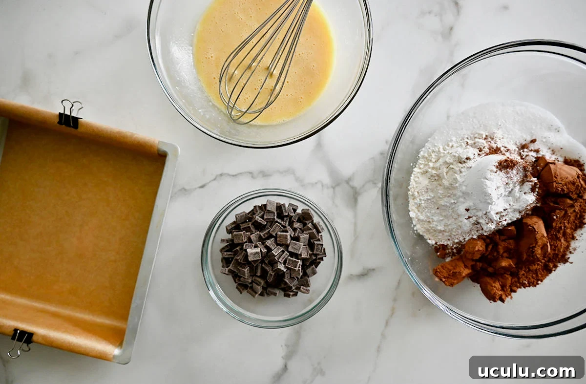 A square baking dish lined with parchment paper next to three glass bowls containing chocolate chunks, flour and cocoa powder, and a whisked egg.