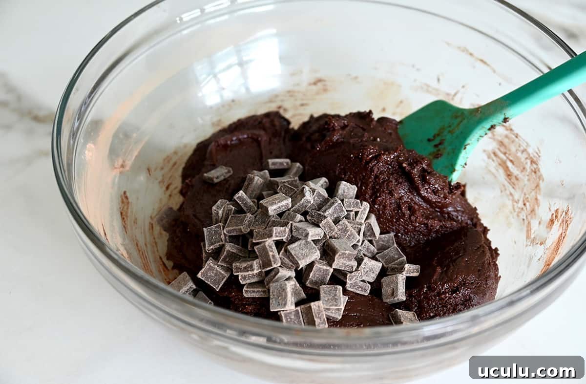 A teal spatula folds chocolate chunks into brownie batter in a glass bowl.