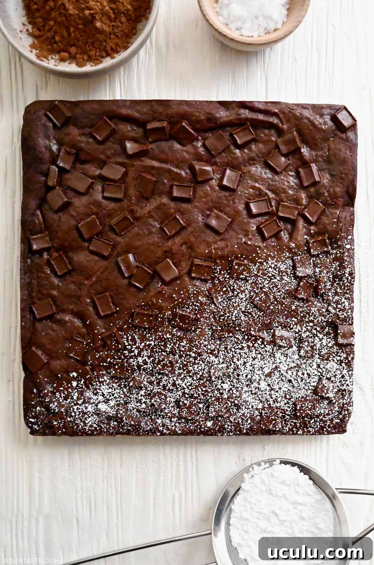 An uncut square brownie with chocolate chunks that's half dusted with powdered sugar.