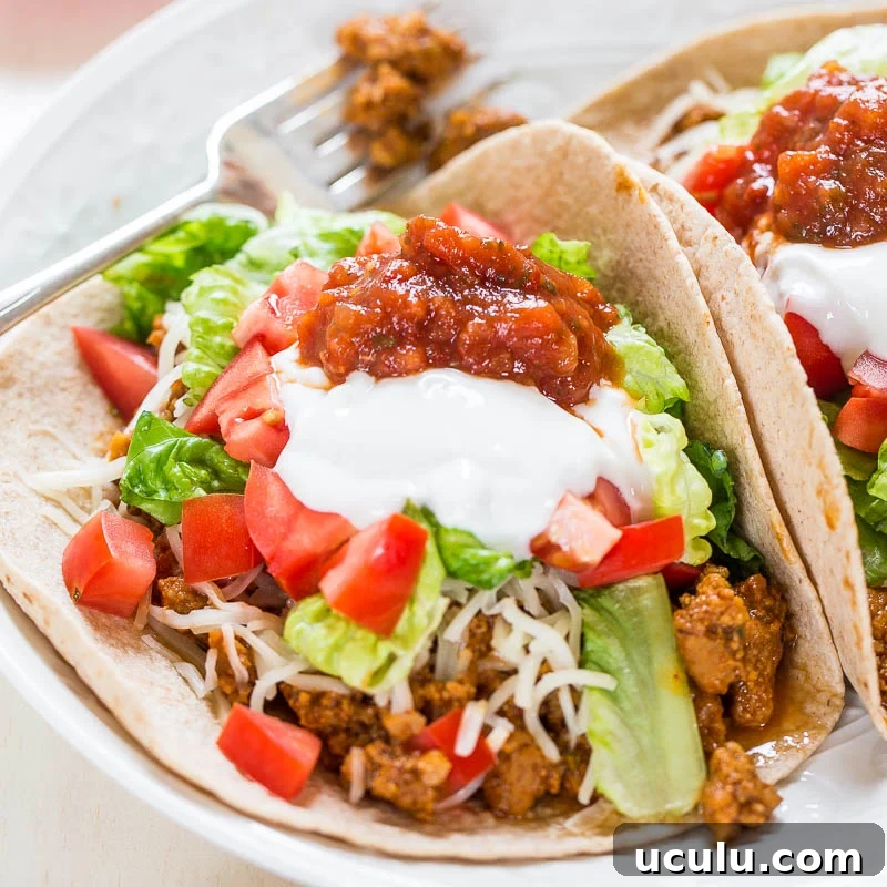 Two tacos with lettuce, tomato, cheese, sour cream, and ground beef on a plate.