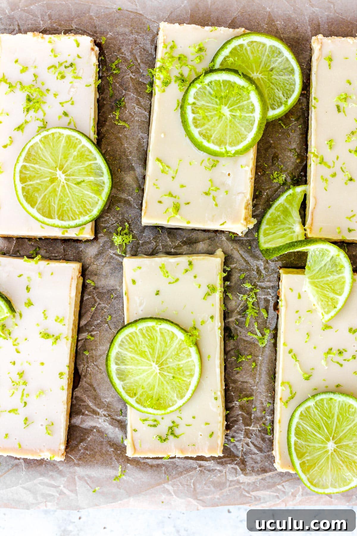Key lime pie bars with fresh lime slices and lime zest on brown parchment paper.