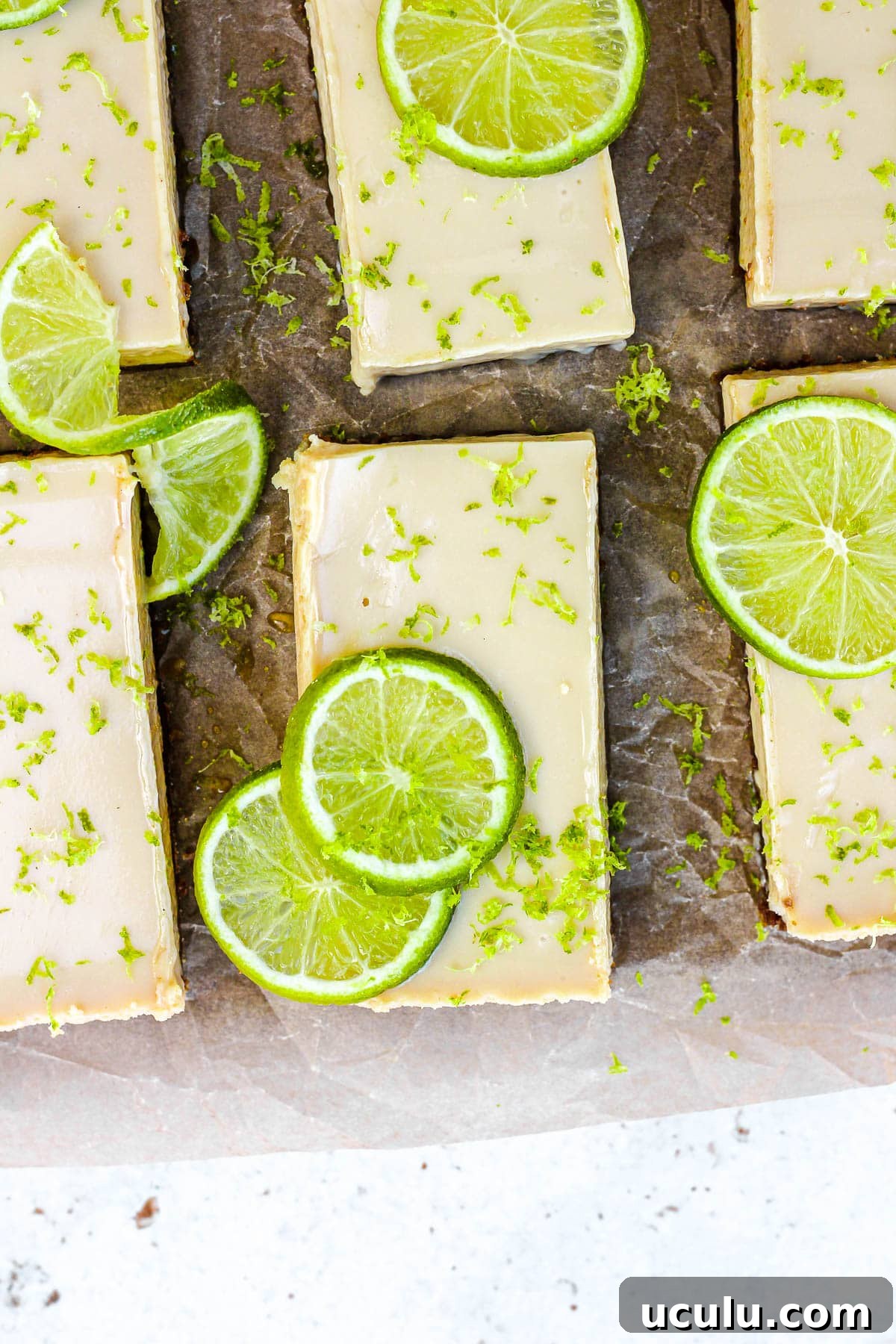Key lime pie bars with fresh lime slices and lime zest on brown parchment paper.