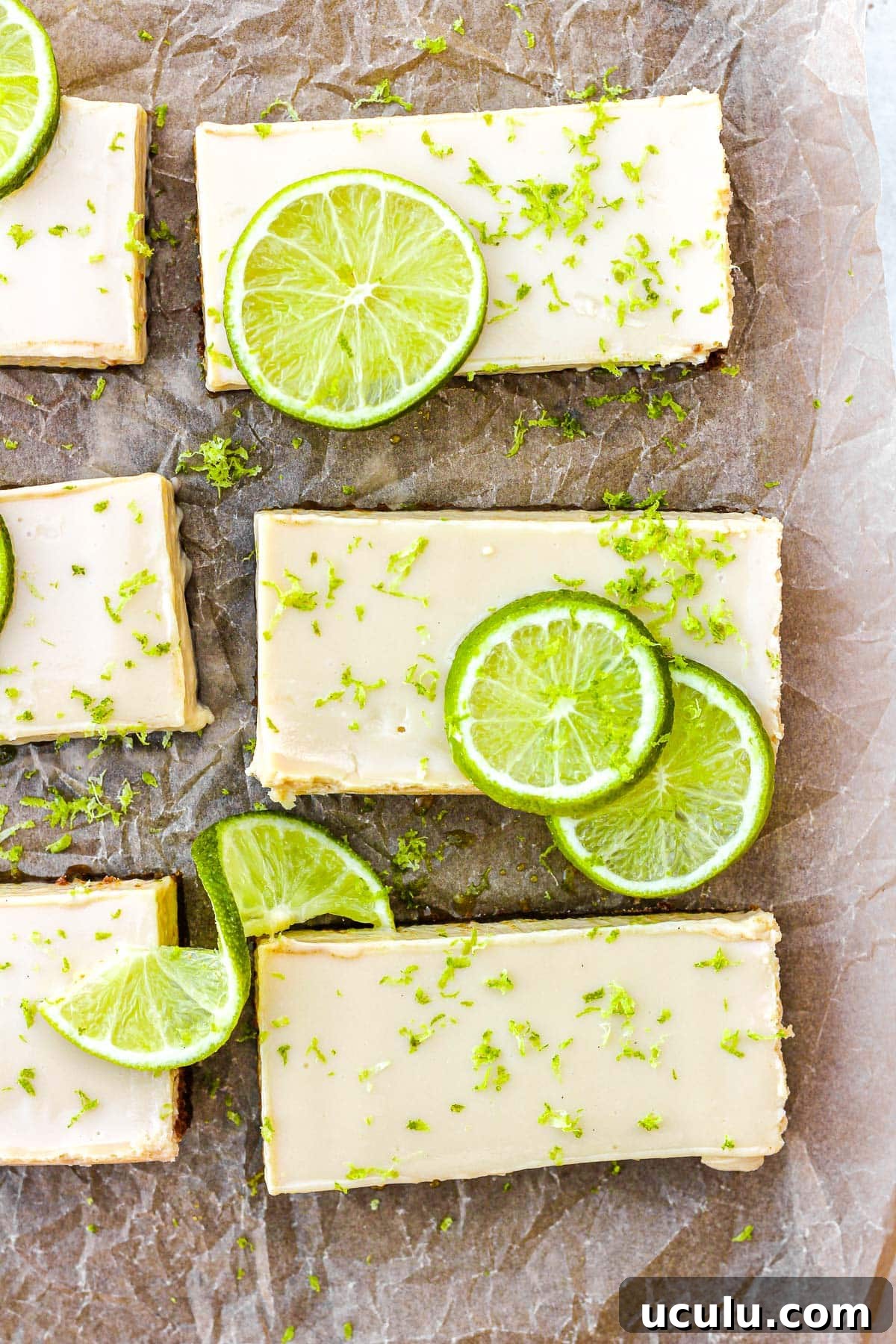 Key lime pie bars with fresh lime slices and lime zest on brown parchment paper.