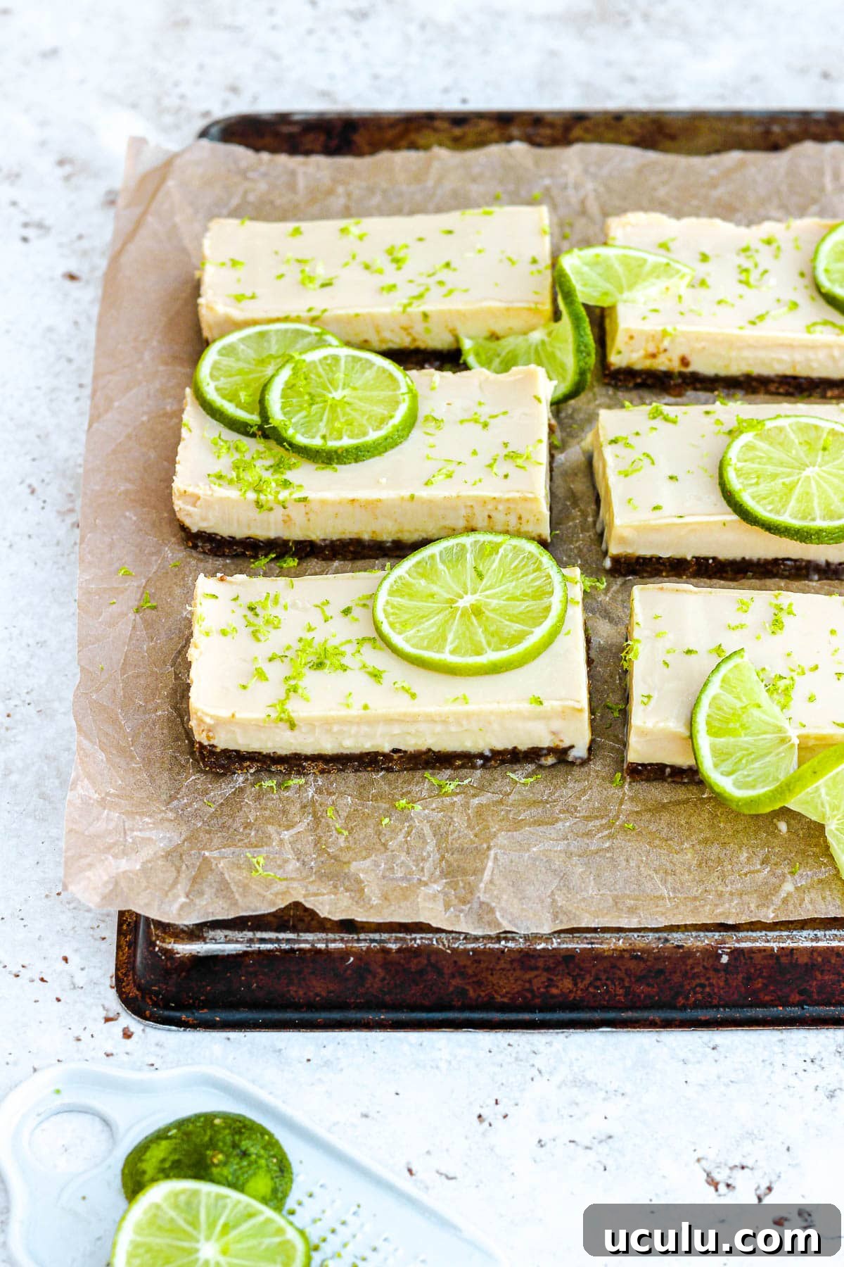 Key lime pie bars with fresh lime slices and lime zest on brown parchment paper.
