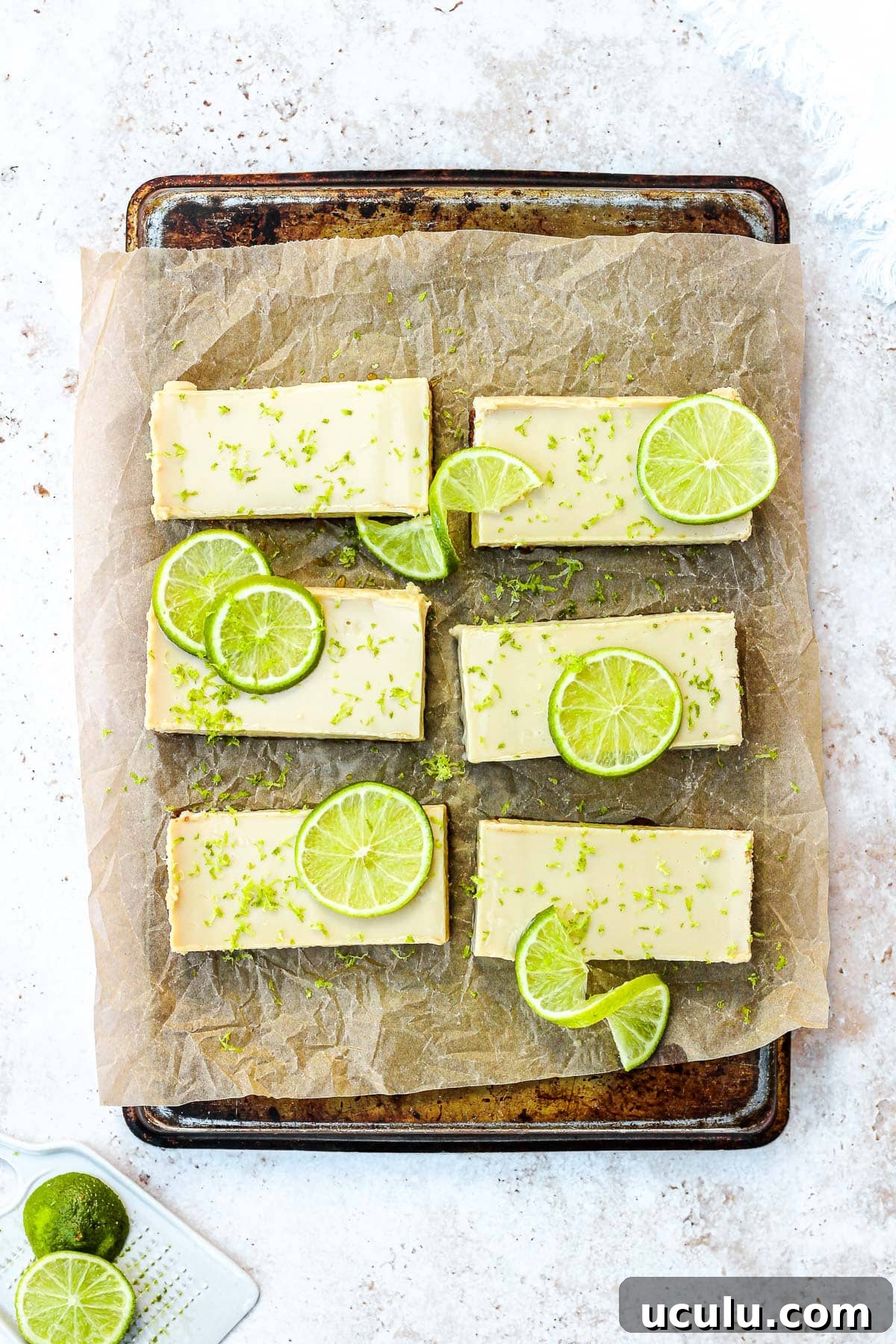 Key lime pie bars with fresh lime slices and lime zest on brown parchment paper.