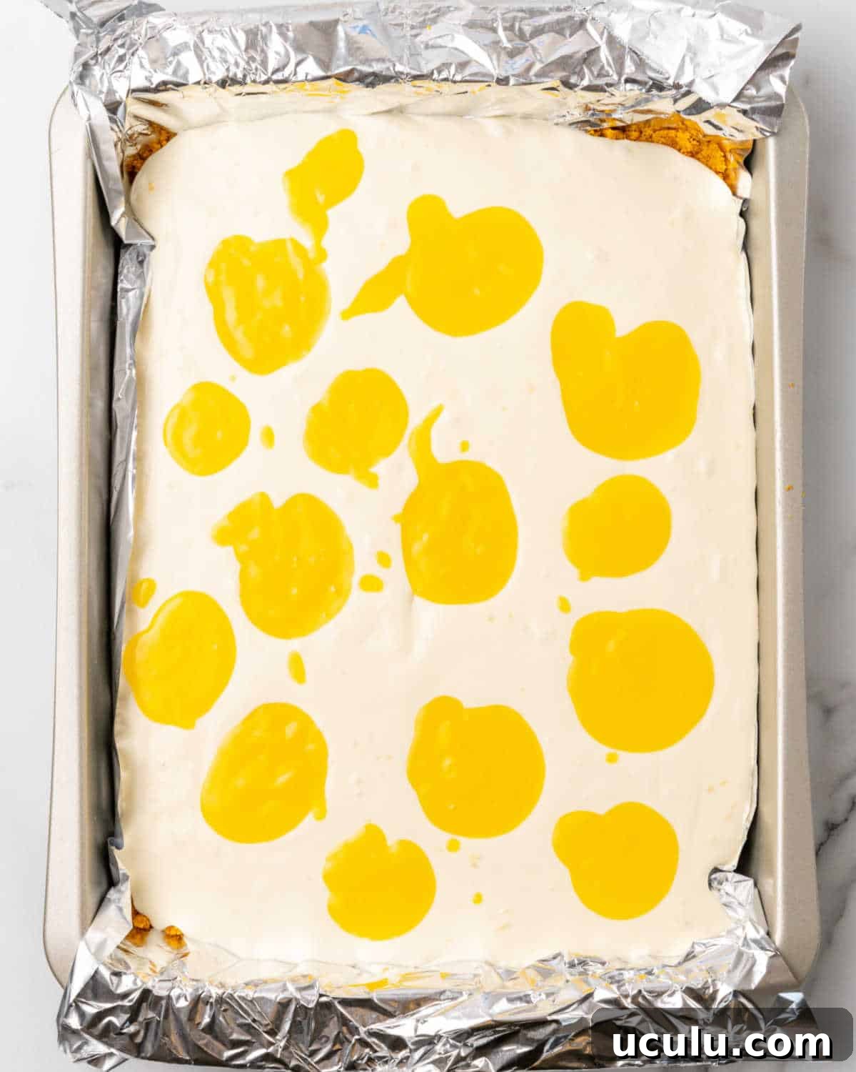 No bake cheesecake batter with mounds of passionfruit curd in a foil-lined rectangular pan on a white surface.